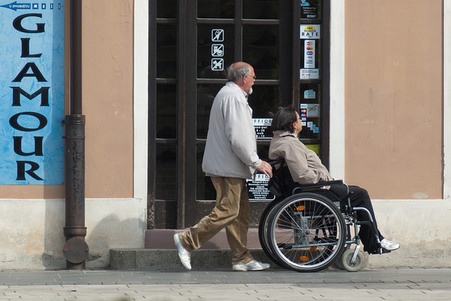 Image is of a man pushing a lady's wheelchair along a footpath. There's a brick building in the background.